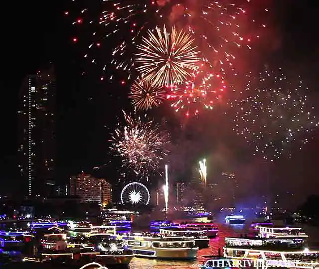 View from the river of widespread, warm-toned golden firework displays illuminating the night over the water, with multiple passenger and riverboat lights reflecting.