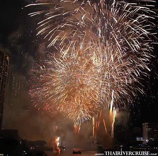 Grand, intricate golden and bronze firework shells create a dense, brilliant display in the night sky over the water, with silhouetted city buildings in the distance.