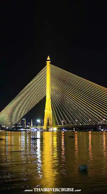 Vertical photograph of the majestic, modern Rama VIII cable-stayed bridge over the Chao Phraya River at night. The bridge and its cables are lit in vibrant gold, and intense gold and yellow light reflections dominate the dark water in the foreground, stretching toward the far bank where city lights are visible. Vertical photograph of the majestic, modern Rama VIII cable-stayed bridge over the Chao Phraya River at night. The bridge and its cables are lit in vibrant gold, and intense gold and yellow light reflections dominate the dark water in the foreground, stretching toward the far bank where city lights are visible.